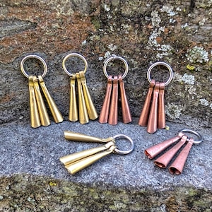 May include: Six sets of three small, cone-shaped bells hanging from silver rings. The bells are made of brass and copper and are arranged in a row on a gray, textured surface.