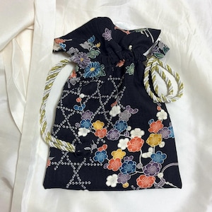 May include: Black drawstring bag with a floral pattern in shades of orange, blue, yellow, and purple. The bag features a geometric design and is accented with gold and white rope drawstrings. The bag is likely made of fabric.