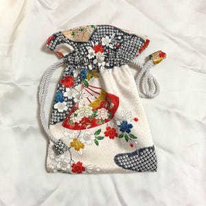 Puede incluir: Pequeña bolsa con cordón y estampado floral. La bolsa presenta un fondo blanco con flores rojas, azules y amarillas, y un diseño de abanico. La parte superior es negra con un patrón de puntos blancos. Tiene un cordón blanco.