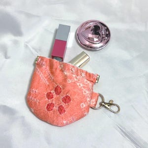 Puede incluir: Un pequeño neceser de color coral con un estampado floral y un cierre dorado. La bolsa contiene un labial rosa, un labial dorado y una polvera rosa con diseño de corazón. Los artículos están sobre una superficie blanca.