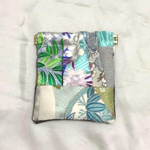 以下が含まれることがあります： 金色の留め金が付いたパッチワークの小銭入れ。緑、青、紫、白の色合いの、花柄や葉柄など、さまざまな生地の模様が特徴です。全体的な美しさは、テクスチャと色のミックスです。