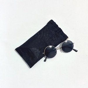 May include: Round sunglasses with thin silver-toned metal frames and dark lenses, positioned next to a rectangular black fabric pouch featuring a subtle, tonal floral jacquard pattern and a small gold-toned metal zipper pull at the top corner.