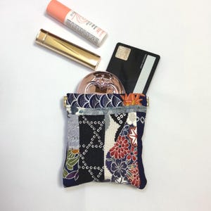 以下が含まれることがあります： 金色の留め金が付いた小さなパッチワーク生地のポーチ。コンパクト、クレジットカード、リップバームのチューブが入っています。ポーチは、ネイビー、黒、グレー、花柄のデザインの模様の生地が特徴です。金色の口紅チューブがポーチの横にあります。