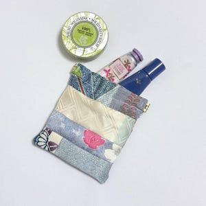 May include: A patchwork fabric pouch in pastel shades, containing a tube of Rose Jasmin hand cream, a blue bottle, and a round tin of Baume Solidarite balm. The pouch is constructed from diverse patterned fabrics.