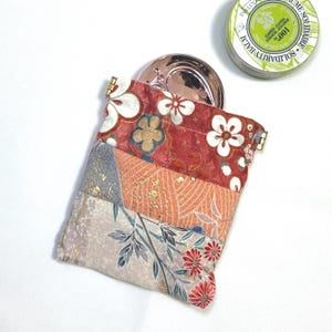 May include: A patchwork fabric pouch with a floral design in red, orange, and cream colors. The pouch is open, revealing a rose gold compact mirror. A green and white tin of balm is also visible.