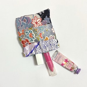 May include: A small, patterned fabric pouch with a gold zipper, holding a white lipstick tube, a pink lip gloss, and a tube of hand cream. The pouch features floral and geometric designs in various colors.