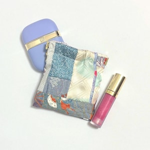 Puede incluir: Un estuche rectangular de color morado claro con una banda dorada, una pequeña bolsa cuadrada de tela de retazos con patrones azules, crema y rojos, y un tubo cilíndrico de brillo labial rosa con tapa dorada sobre fondo blanco.