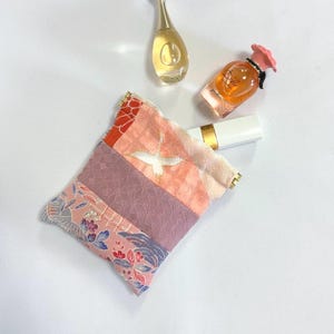 Può includere: Un piccolo astuccio in tessuto patchwork con un disegno di uccello, nei toni del rosa, arancione e viola. L'astuccio è accanto a un rossetto bianco e due flaconi di profumo. L'astuccio ha dettagli dorati.