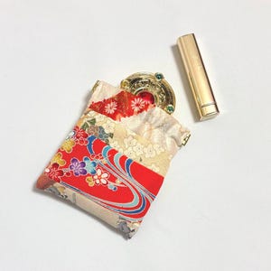Peut inclure: Une petite pochette en tissu ornée d'un motif floral rouge, blanc et doré avec des lignes bleues tourbillonnantes. À l'intérieur se trouve un poudrier rond doré avec un centre rouge et des accents verts, accompagné d'un tube de rouge à lèvres.