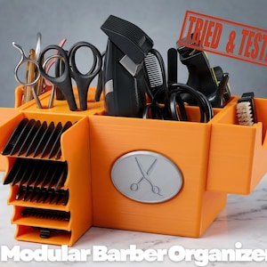 May include: An orange modular barber organiser with multiple compartments. It holds various barber tools, including scissors, clippers, and combs. The organiser has a silver emblem with crossed scissors and the text "Modular Barber Organizer". A red stamp reads "Tried & Tested".
