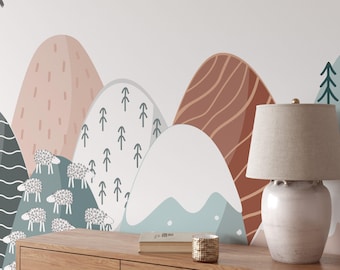 May include: A white ceramic table lamp with a beige linen shade sits on a wooden table in front of a wall mural depicting a whimsical landscape of mountains, trees, and sheep. The mountains are painted in shades of pink, brown, and blue.