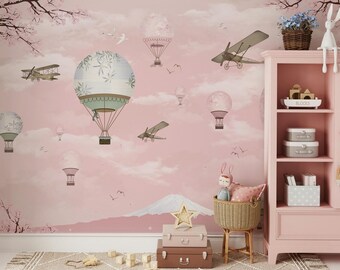 Könnte beinhalten: Ein rosa Kinderzimmer-Wandbild mit einem verspielten Design mit Heißluftballons, Flugzeugen und Kirschblüten. Das Wandbild wird durch ein weißes Holzregalsystem mit hellrosa Finish akzentuiert.