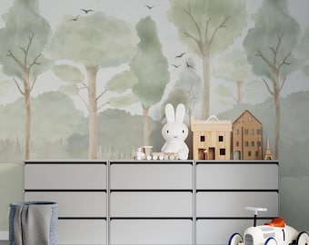 May include: A white toy bunny sits on top of a set of gray drawers with a toy train and two wooden houses. The drawers are in front of a wall with a watercolor mural of a forest with green trees and birds.