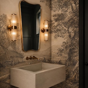 May include: A contemporary bathroom with a rectangular concrete sink, a gold-edged mirror, and two wall lights with cylindrical glass shades. The wallpaper shows a sepia-toned landscape scene.