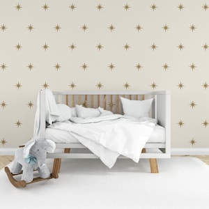 Puede incluir: Una cuna blanca con un colchón blanco y ropa de cama blanca. La cuna está frente a una pared con un patrón de estrellas doradas. Hay un caballo de balancín en primer plano.