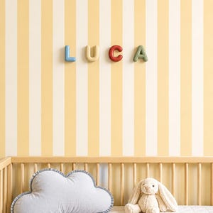 Pode incluir: Uma cena de berçário com o nome "LUCA" escrito em letras 3D coloridas em uma parede listrada amarela e branca. Uma almofada em forma de nuvem e um coelhinho de pelúcia estão em um berço de madeira.