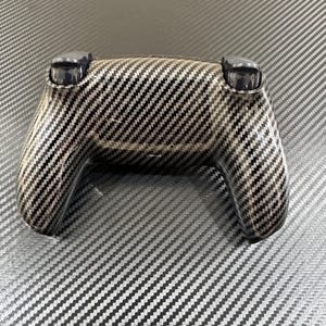 May include: A black and silver controller with a carbon fiber pattern. The controller has two black joysticks and a textured surface. The design is a diagonal weave pattern, giving it a modern and sleek appearance.