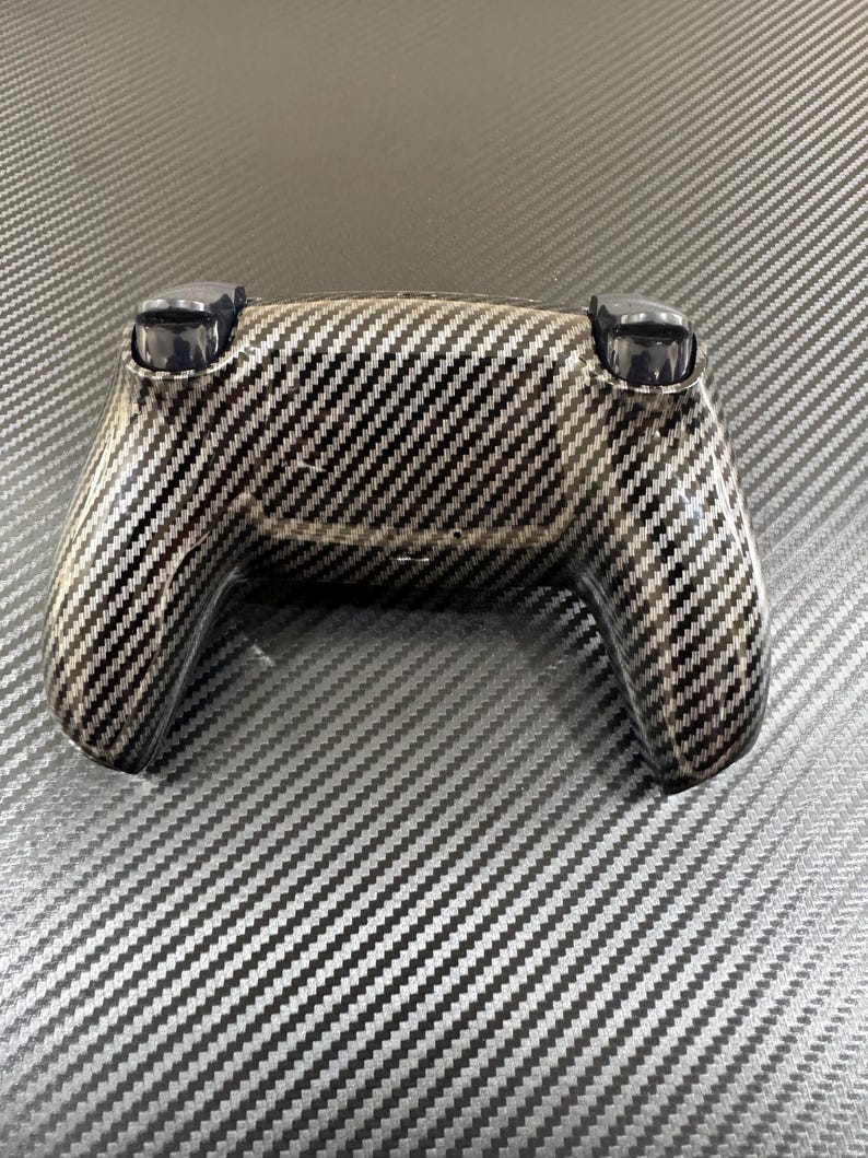 May include: A black and silver controller with a carbon fiber pattern. The controller has two black joysticks and a textured surface. The design is a diagonal weave pattern, giving it a modern and sleek appearance.