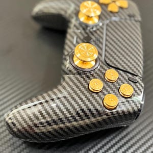 May include: A black and gray carbon fiber gaming controller with gold-colored buttons and accents. The controller has a textured surface and a sleek design, featuring a directional pad and multiple buttons.