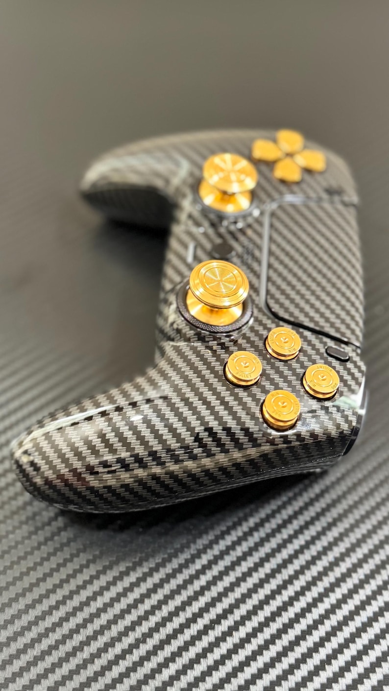 May include: A black and gray carbon fiber gaming controller with gold-colored buttons and accents. The controller has a textured surface and a sleek design, featuring a directional pad and multiple buttons.