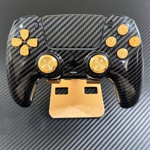 May include: A black and gold gaming controller with a carbon fiber design. The controller features gold-colored buttons and a gold-colored stand. The directional pad and buttons are gold. The controller is designed for gaming.