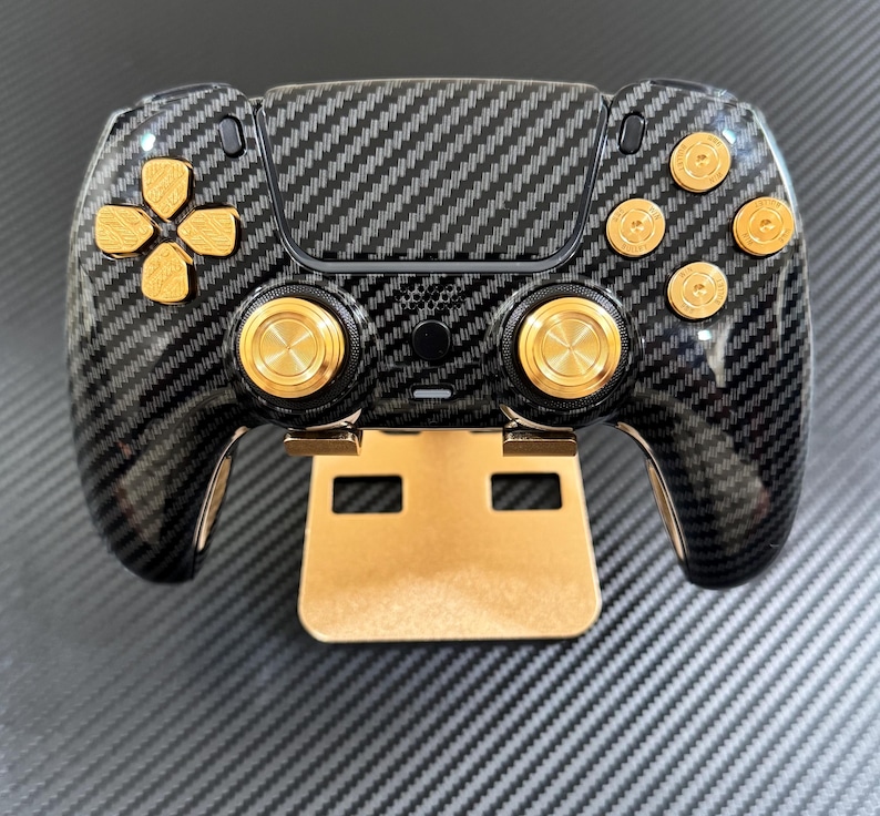 May include: A black and gold gaming controller with a carbon fiber design. The controller features gold-colored buttons and a gold-colored stand. The directional pad and buttons are gold. The controller is designed for gaming.