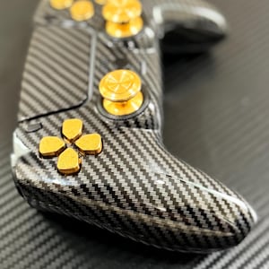 May include: A gaming controller with a carbon fiber design and gold-colored buttons. The controller has a sleek, ergonomic shape with a directional pad, buttons, and analog sticks. The buttons are gold-colored and the controller is black and gray.