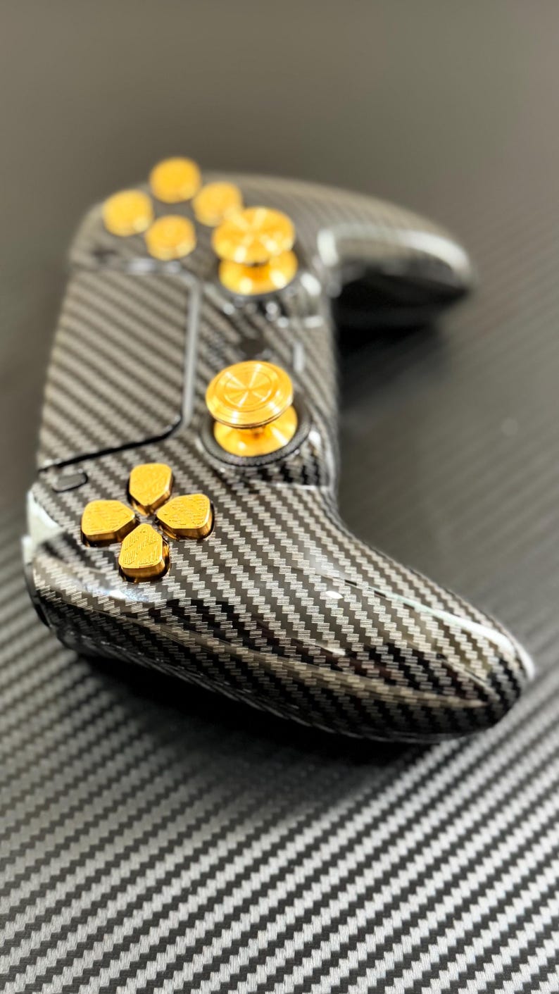 May include: A gaming controller with a carbon fiber design and gold-colored buttons. The controller has a sleek, ergonomic shape with a directional pad, buttons, and analog sticks. The buttons are gold-colored and the controller is black and gray.