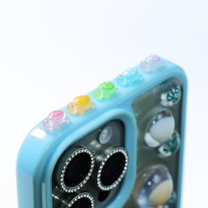 May include: A light blue phone case featuring a row of small, translucent, multi-colored turtle figurines along the top edge. The camera lens area is surrounded by a silver-toned border with embedded clear crystals, and the back has clear accents.