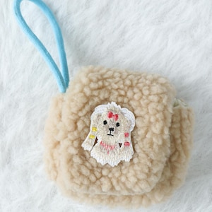 Könnte beinhalten: Kleine, beige, flauschige Tasche mit hellblauem Kordelgriff. Die Tasche hat einen gestickten Aufnäher eines Cartoon-Hundes mit roter Schleife und bunten Akzenten. Die Tasche ist aus einem weichen, strukturierten Material.