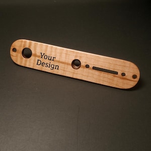 May include: A light-colored wooden guitar part with the text "Your Design" engraved on it. The part has several holes and slots, and is likely a control plate or similar component. The wood grain is visible.