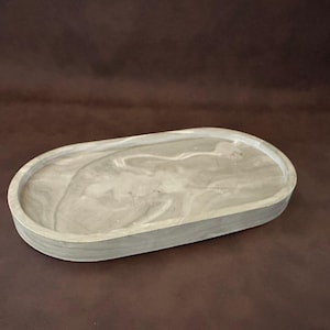 Puede incluir: Bandeja ovalada de hormigón con diseño veteado gris claro. La bandeja tiene un borde elevado y una superficie lisa. El color neutro de la bandeja la hace adecuada para diversos usos. Mide unos 25 cm de largo.