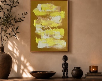 May include: Abstract art piece with a gold frame. The artwork features layered brushstrokes of yellow, white, and beige on a mustard-colored background. The painting hangs above a wooden cabinet with decorative items, including a wooden bowl, a small statue, and a candle.