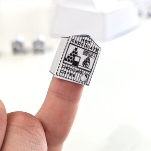 May include: A miniature, black and white paper model of a hot chocolate and coffee shop. The shop features the words "HOT CHOCOLATE" and "CHOCOLATE COFFEE" in a vertical and horizontal layout. A small illustration of a person is visible.
