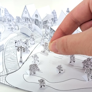 May include: A black and white paper winter village diorama with pop-up houses, trees, and figures. A hand is placing a small paper figure of a person on a snowboard. The scene includes a bridge, street lamps, and other winter-themed elements.