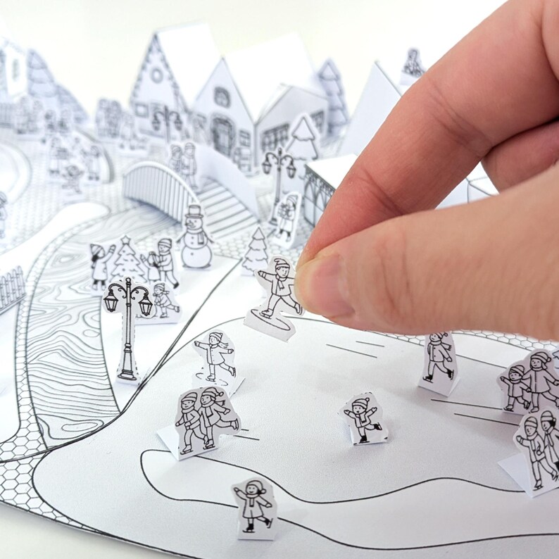 May include: A black and white paper winter village diorama with pop-up houses, trees, and figures. A hand is placing a small paper figure of a person on a snowboard. The scene includes a bridge, street lamps, and other winter-themed elements.
