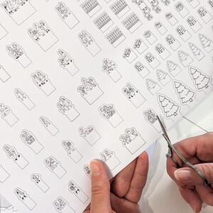 May include: A sheet of white paper with black line art illustrations of Christmas-themed figures and objects. A pair of scissors is cutting out one of the illustrations. The illustrations include Christmas trees, people, and gift boxes.
