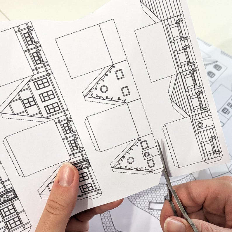 May include: A person is using scissors to cut out a paper house template. The template is printed on white paper and features detailed line drawings of windows, doors, and architectural elements. The design includes dotted lines for folding and cutting.