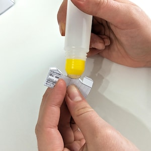 May include: A clear glue bottle dispensing yellow glue onto a small, black and white paper model. The model appears to be a building with detailed architectural elements. The image shows the process of assembling a paper craft project.