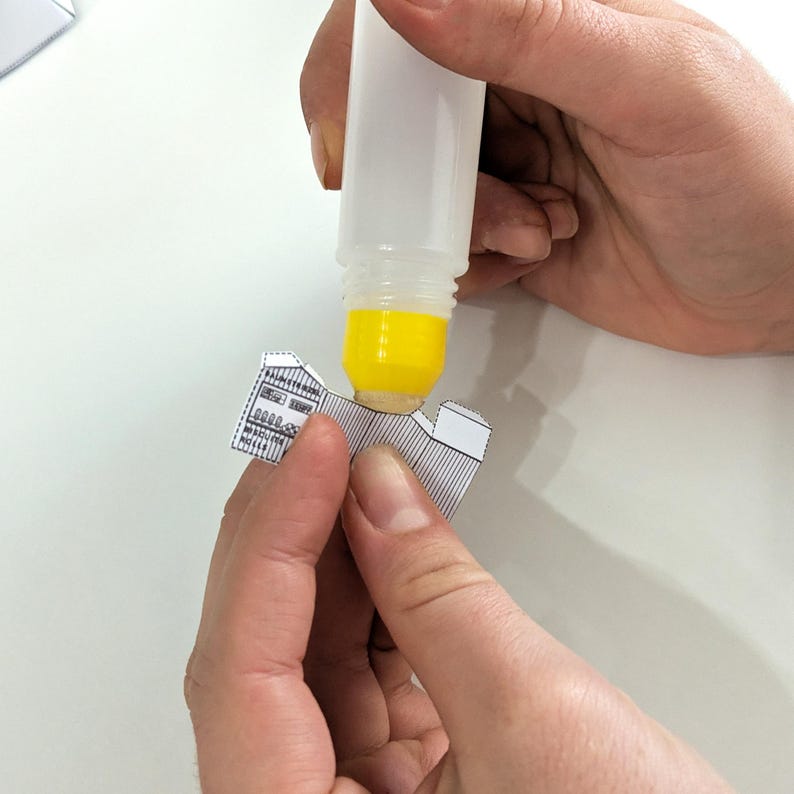 May include: A clear glue bottle dispensing yellow glue onto a small, black and white paper model. The model appears to be a building with detailed architectural elements. The image shows the process of assembling a paper craft project.