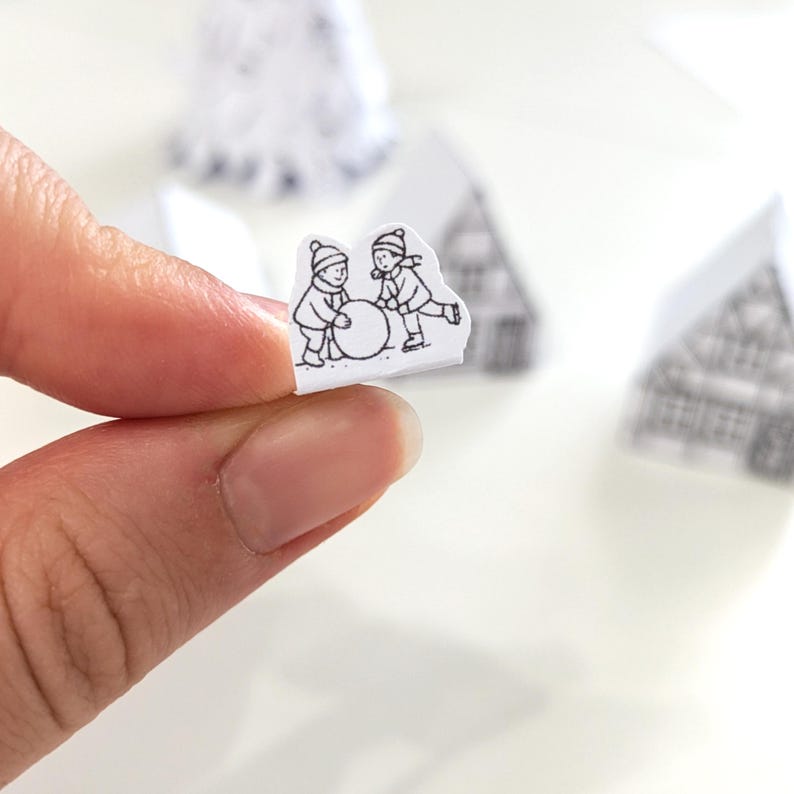 May include: A small, black and white illustration of two children building a snowman. The image is held between fingers, with other miniature paper houses in the background. The scene evokes a winter theme.