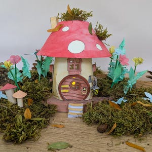 May include: A miniature mushroom-shaped house with a red roof featuring white spots, a rounded brown door, and a small lantern. The structure is surrounded by green moss, paper flowers, and small brown leaves, creating a detailed woodland scene.