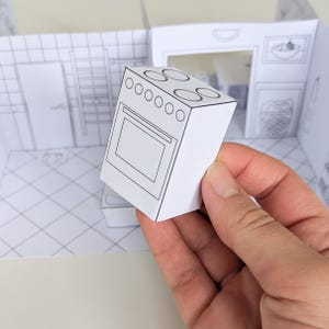 May include: A small, three-dimensional paper cutout of a kitchen stove with black line drawings on white paper. The stove features four circular burners on top and a rectangular oven door. It is positioned in front of a flat, printed paper room backdrop.