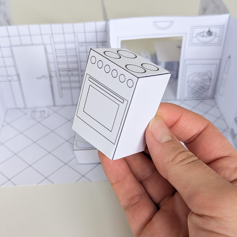 May include: A small, three-dimensional paper cutout of a kitchen stove with black line drawings on white paper. The stove features four circular burners on top and a rectangular oven door. It is positioned in front of a flat, printed paper room backdrop.