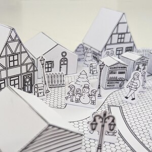 May include: A black and white paper diorama of a winter village scene. The scene includes houses, shops, a snowman, children, and street lamps. The diorama is made of folded paper and has a three-dimensional effect.