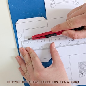 May include: A red craft knife is positioned against a clear plastic ruler measuring a white paper template with printed line drawings. The ruler shows markings in centimeters, with the blade aligned at the zero mark on the paper surface.