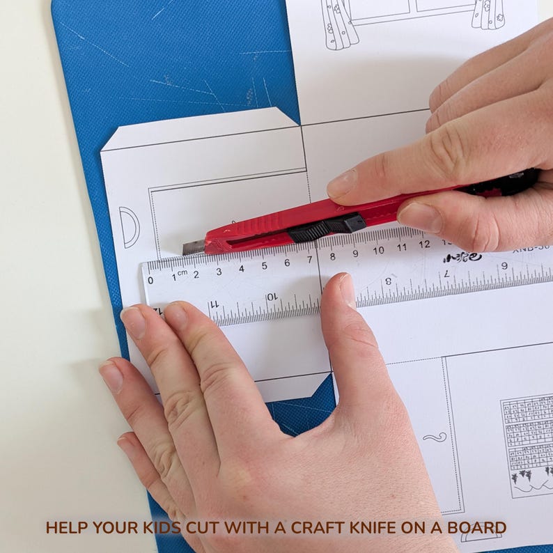 May include: A red craft knife is positioned against a clear plastic ruler measuring a white paper template with printed line drawings. The ruler shows markings in centimeters, with the blade aligned at the zero mark on the paper surface.