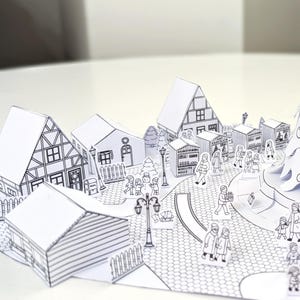 May include: A detailed, black and white papercraft village scene. The pop-up design features buildings, a Christmas tree, and figures of people. The scene includes a street, lampposts, and a snowman, creating a festive holiday atmosphere.
