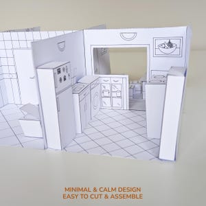 May include: A white paper craft model of a kitchen interior featuring black line drawings of a refrigerator, sink, washing machine, cabinets, and a bowl of fruit. The structure includes a tiled floor pattern and walls with cut-out details.