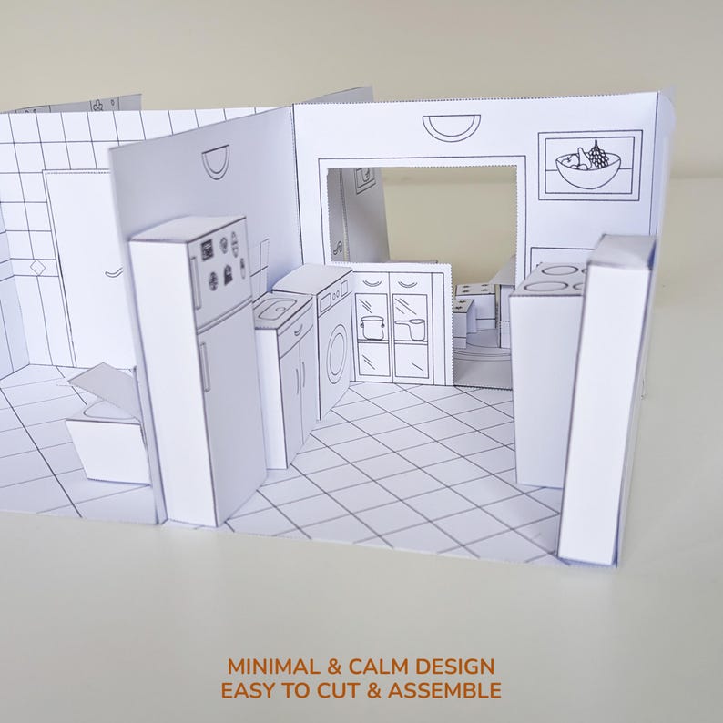 May include: A white paper craft model of a kitchen interior featuring black line drawings of a refrigerator, sink, washing machine, cabinets, and a bowl of fruit. The structure includes a tiled floor pattern and walls with cut-out details.